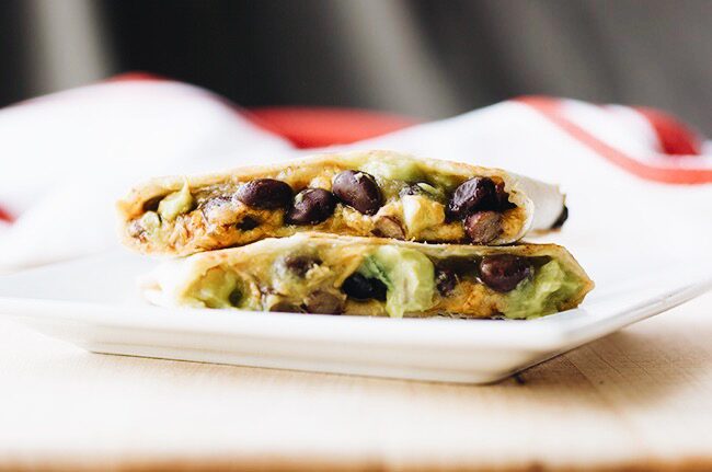 Close-up cross-section view of a collard greens and black bean quesadilla showing the melted cheese, vibrant green collards, and black beans, with a small bowl of salsa verde in the background