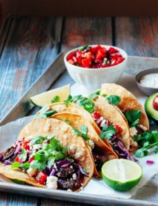 Perfectly grilled carne asada tacos on warm corn tortillas with fresh cilantro, diced onions, and lime wedges, beautifully plated on a rustic wooden board