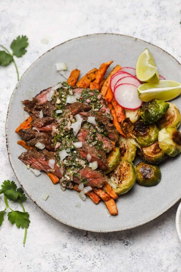 Raw marinated skirt steak on a cutting board next to vibrant marinade ingredients including lime halves, garlic, and fresh herbs