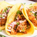 Chicken tinga served on warm tortillas with fresh cilantro, diced onions, and lime wedges, showing the rich reddish-brown sauce coating tender shredded chicken