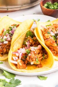 Chicken tinga served on warm tortillas with fresh cilantro, diced onions, and lime wedges, showing the rich reddish-brown sauce coating tender shredded chicken