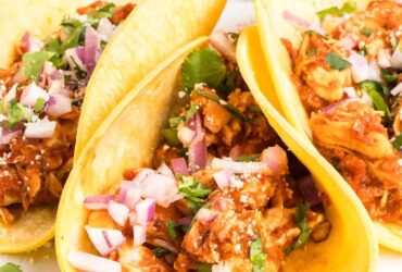Chicken tinga served on warm tortillas with fresh cilantro, diced onions, and lime wedges, showing the rich reddish-brown sauce coating tender shredded chicken