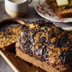 Chocolate chunk banana bread sliced on wooden cutting board with chocolate chunks visible throughout, golden brown exterior, served with coffee cup in background