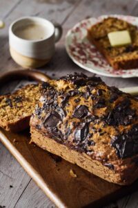 Chocolate chunk banana bread sliced on wooden cutting board with chocolate chunks visible throughout, golden brown exterior, served with coffee cup in background