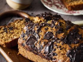 Chocolate chunk banana bread sliced on wooden cutting board with chocolate chunks visible throughout, golden brown exterior, served with coffee cup in background