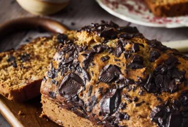 Chocolate chunk banana bread sliced on wooden cutting board with chocolate chunks visible throughout, golden brown exterior, served with coffee cup in background