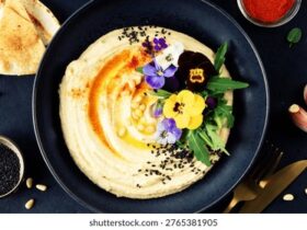 Perfectly smooth creamy hummus in a wide shallow bowl, drizzled with golden olive oil and garnished with paprika, chickpeas, and fresh herbs, photographed from above on a rustic wooden table