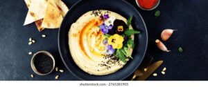 Perfectly smooth creamy hummus in a wide shallow bowl, drizzled with golden olive oil and garnished with paprika, chickpeas, and fresh herbs, photographed from above on a rustic wooden table