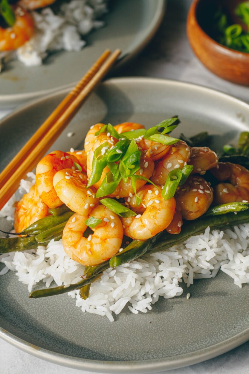 close up of glazed honey garlic shrimp served over white rice with chopsticks and garnished with sliced green onions