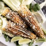 Charred Mexican street corn on wooden skewers drizzled with creamy white sauce, sprinkled with crumbled cotija cheese and bright red chili powder, garnished with lime wedges on a rustic wooden board