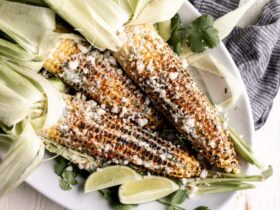 Charred Mexican street corn on wooden skewers drizzled with creamy white sauce, sprinkled with crumbled cotija cheese and bright red chili powder, garnished with lime wedges on a rustic wooden board