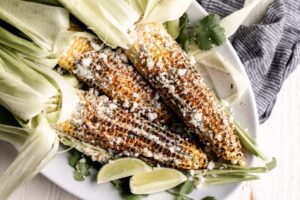 Charred Mexican street corn on wooden skewers drizzled with creamy white sauce, sprinkled with crumbled cotija cheese and bright red chili powder, garnished with lime wedges on a rustic wooden board
