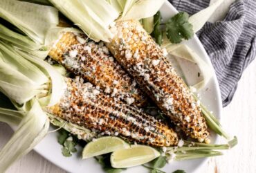 Charred Mexican street corn on wooden skewers drizzled with creamy white sauce, sprinkled with crumbled cotija cheese and bright red chili powder, garnished with lime wedges on a rustic wooden board