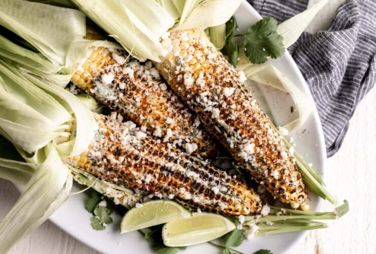 Charred Mexican street corn on wooden skewers drizzled with creamy white sauce, sprinkled with crumbled cotija cheese and bright red chili powder, garnished with lime wedges on a rustic wooden board