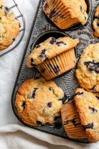 Beautiful golden-brown blueberry muffins with towering tops in a muffin tin, some cooling on a wire rack, with fresh blueberries scattered around