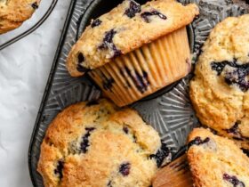 Beautiful golden-brown blueberry muffins with towering tops in a muffin tin, some cooling on a wire rack, with fresh blueberries scattered around
