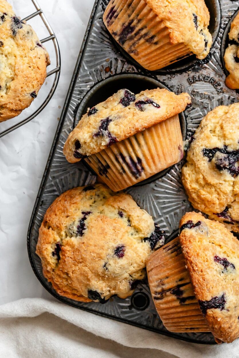 Beautiful golden-brown blueberry muffins with towering tops in a muffin tin, some cooling on a wire rack, with fresh blueberries scattered around