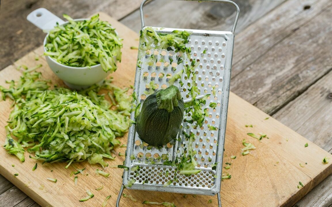 hands grating fresh zucchini on box grater with pile of shredded zucchini