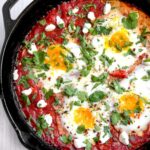 Shakshuka in cast iron skillet with golden runny egg yolks and fresh herbs, overhead shot with warm lighting