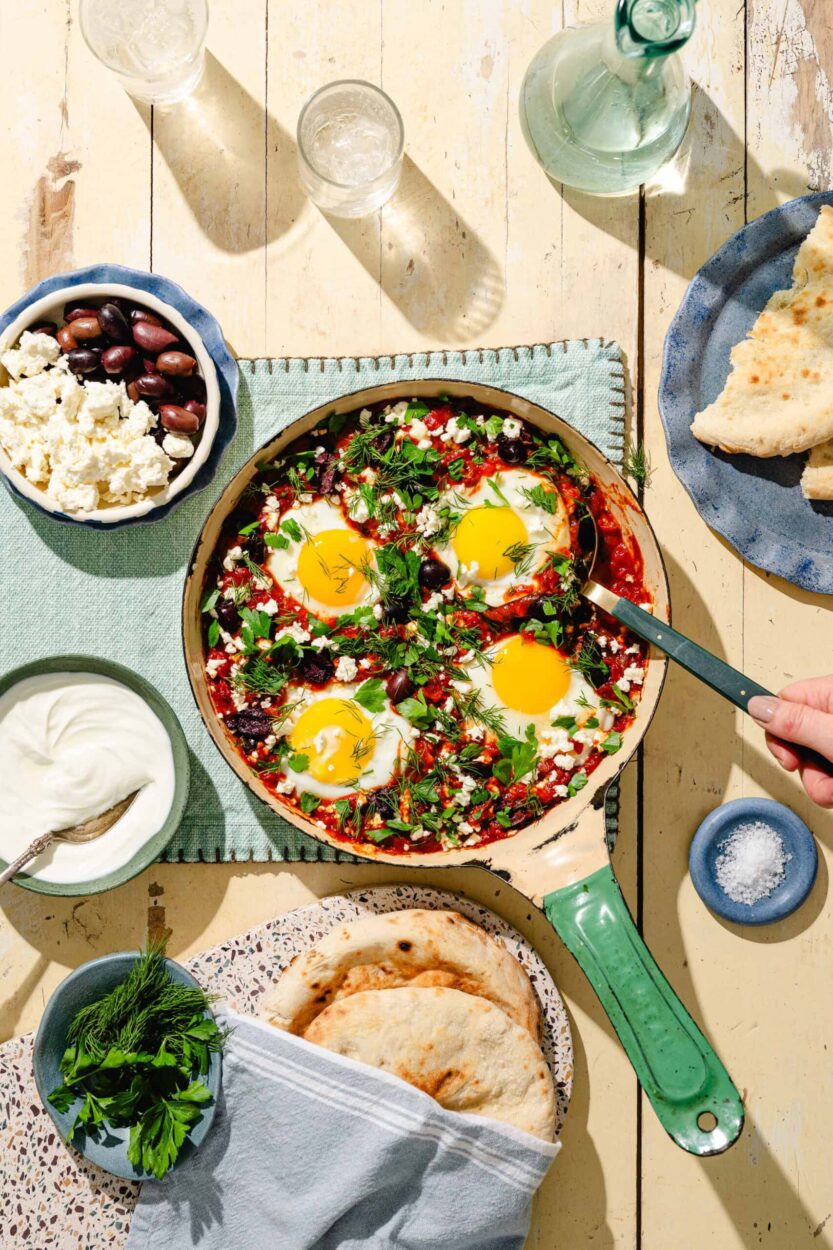 Close-up of shakshuka sauce bubbling in skillet with spices and fresh herbs scattered around