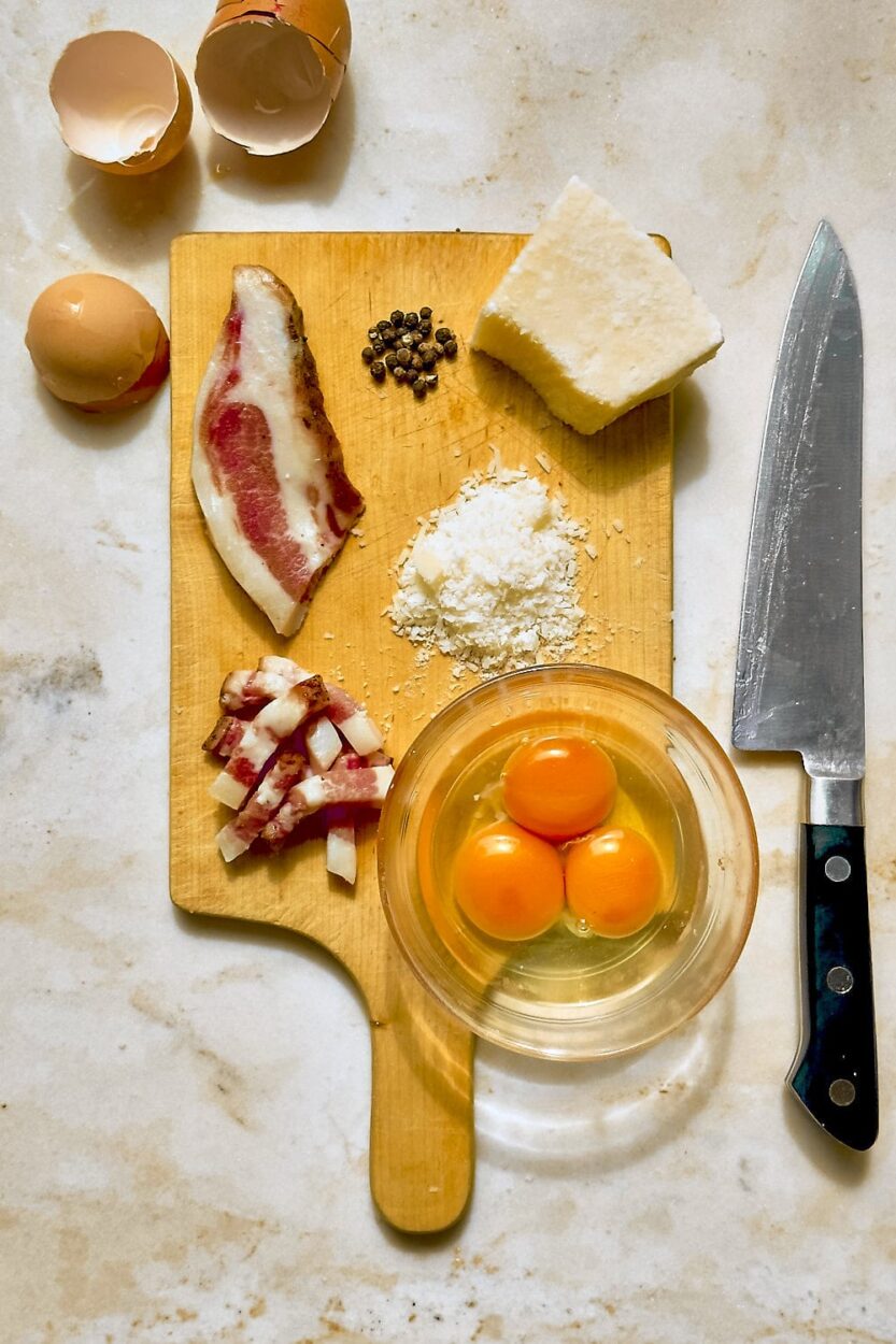Raw guanciale being diced on a wooden cutting board next to whole eggs and grated pecorino romano cheese