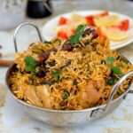 Overhead shot of golden chicken biryani in a heavy-bottomed pot with visible saffron threads, garnished with fresh cilantro and crispy onions, with a serving spoon ready to dig in