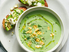 Elegant cold cucumber avocado soup served in a white ceramic bowl with a swirl of cream, fresh herbs, and a drizzle of olive oil, photographed from above on a marble surface
