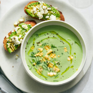 Elegant cold cucumber avocado soup served in a white ceramic bowl with a swirl of cream, fresh herbs, and a drizzle of olive oil, photographed from above on a marble surface