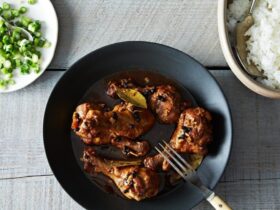 Filipino chicken adobo served in a rustic bowl with glossy dark sauce coating tender chicken pieces, garnished with fresh scallions, with steamed rice on the side