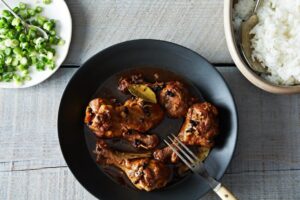 Filipino chicken adobo served in a rustic bowl with glossy dark sauce coating tender chicken pieces, garnished with fresh scallions, with steamed rice on the side