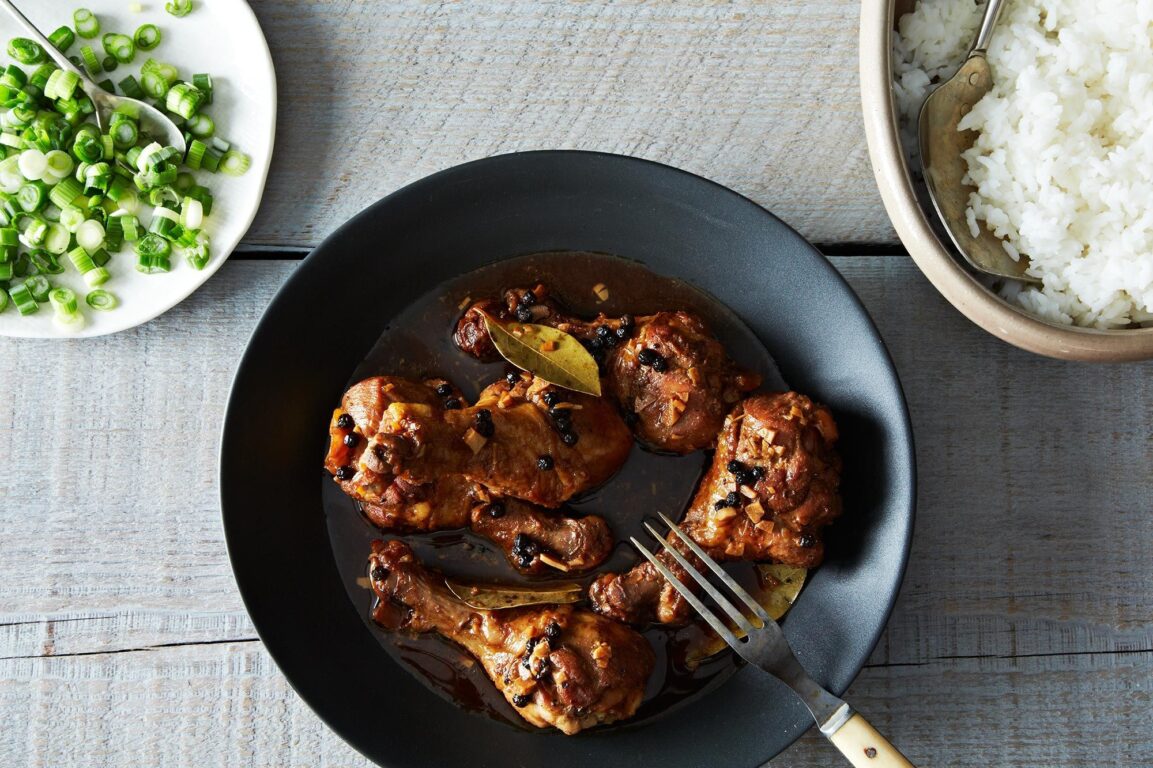 Filipino chicken adobo served in a rustic bowl with glossy dark sauce coating tender chicken pieces, garnished with fresh scallions, with steamed rice on the side