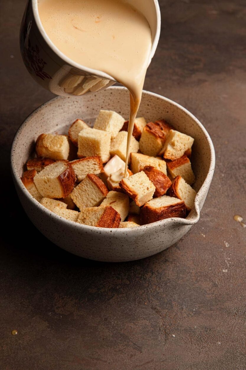 Overhead shot of brioche bread cubes in baking dish being covered with creamy custard mixture from a glass measuring cup
