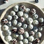 A beautiful overhead shot of frozen yogurt covered blueberries arranged on a white marble surface, glistening with a creamy yogurt shell, scattered with fresh blueberries and mint leaves for garnish