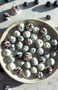 A beautiful overhead shot of frozen yogurt covered blueberries arranged on a white marble surface, glistening with a creamy yogurt shell, scattered with fresh blueberries and mint leaves for garnish