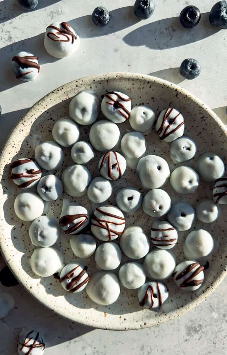 A beautiful overhead shot of frozen yogurt covered blueberries arranged on a white marble surface, glistening with a creamy yogurt shell, scattered with fresh blueberries and mint leaves for garnish