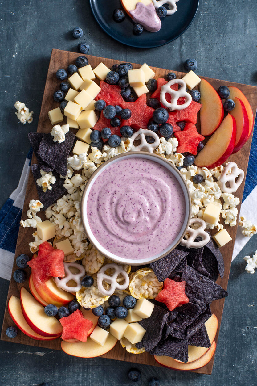 A step-by-step flat lay showing fresh blueberries in a bowl, a cup of creamy Greek yogurt with honey being drizzled in, toothpicks, and a parchment-lined baking sheet ready for freezing