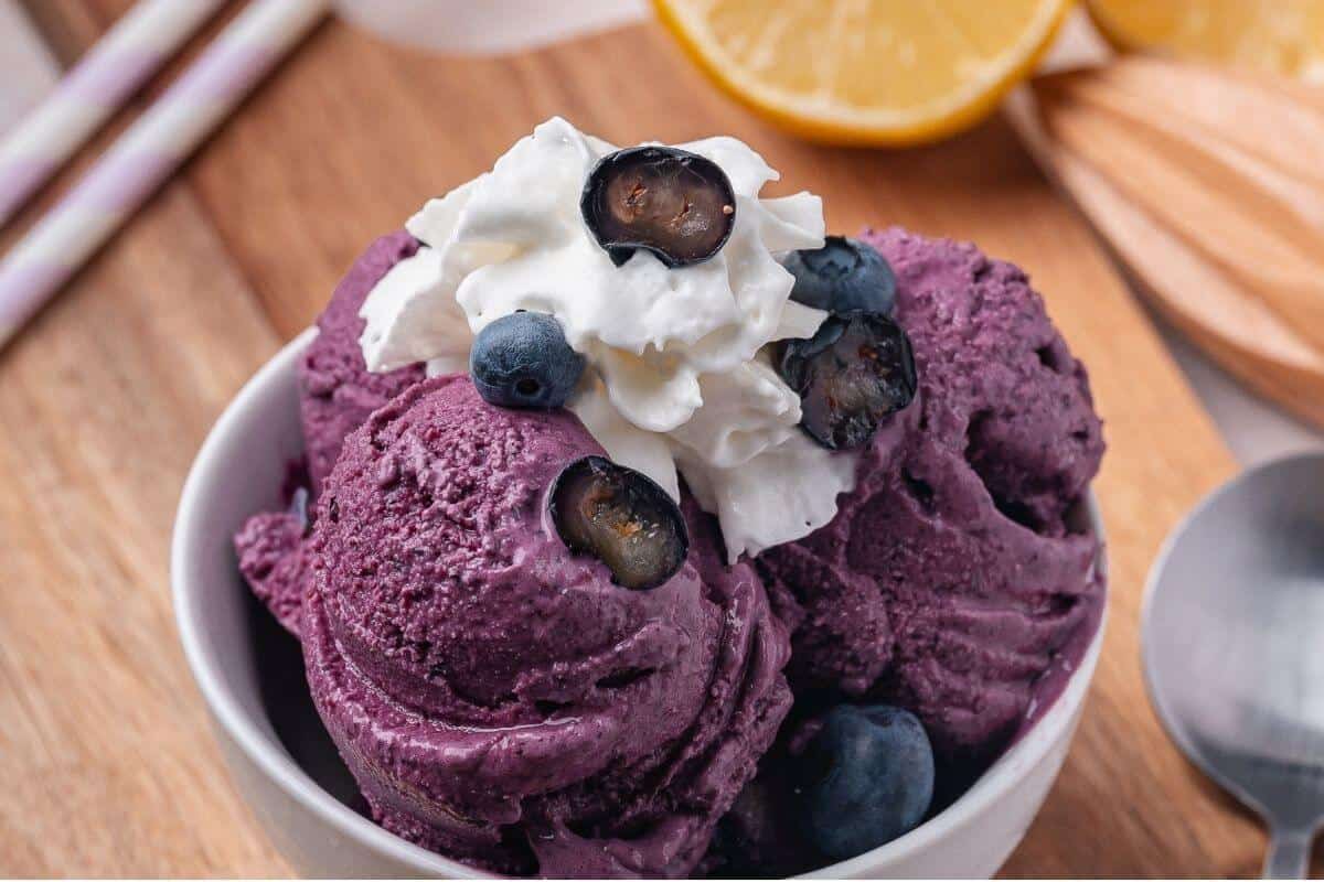 A close-up macro shot of frozen yogurt covered blueberries piled in a small white ceramic bowl, the creamy yogurt shells catching the light, with a single berry cut in half to reveal the vivid purple interior