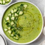 A beautifully plated bowl of gazpacho verde with avocado, garnished with sliced cucumber, a drizzle of olive oil, microgreens, and a swirl of cream on a white ceramic bowl against a light linen backdrop