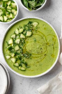 A beautifully plated bowl of gazpacho verde with avocado, garnished with sliced cucumber, a drizzle of olive oil, microgreens, and a swirl of cream on a white ceramic bowl against a light linen backdrop