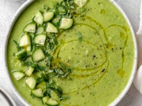 A beautifully plated bowl of gazpacho verde with avocado, garnished with sliced cucumber, a drizzle of olive oil, microgreens, and a swirl of cream on a white ceramic bowl against a light linen backdrop