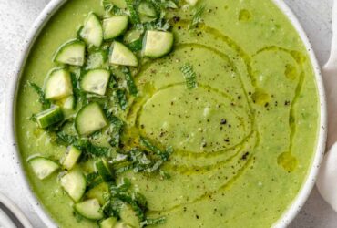A beautifully plated bowl of gazpacho verde with avocado, garnished with sliced cucumber, a drizzle of olive oil, microgreens, and a swirl of cream on a white ceramic bowl against a light linen backdrop