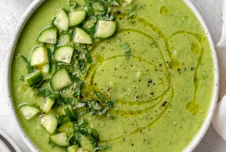 A beautifully plated bowl of gazpacho verde with avocado, garnished with sliced cucumber, a drizzle of olive oil, microgreens, and a swirl of cream on a white ceramic bowl against a light linen backdrop