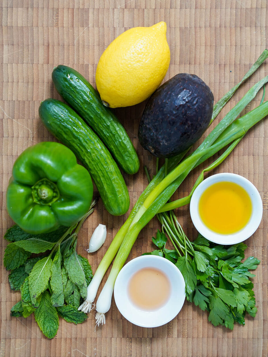 Flat-lay ingredients shot showing halved avocados, sliced English cucumber, green bell pepper, garlic cloves, fresh herbs, a bottle of sherry vinegar, and extra-virgin olive oil arranged on a pale green marble surface