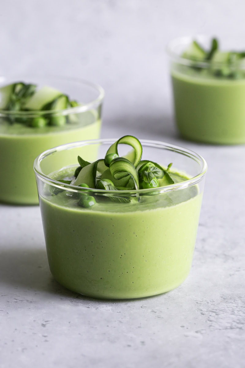 Close-up serving shot of two chilled white bowls of gazpacho verde with avocado garnished with thin cucumber ribbons, a swirl of olive oil, fresh dill fronds, and cracked black pepper, with a linen napkin and silver spoons beside them on a wooden table
