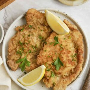 Golden brown German schnitzel on an elegant white plate, garnished with fresh lemon wedges and parsley, with a side of crispy potatoes