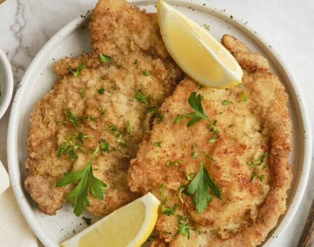 Golden brown German schnitzel on an elegant white plate, garnished with fresh lemon wedges and parsley, with a side of crispy potatoes