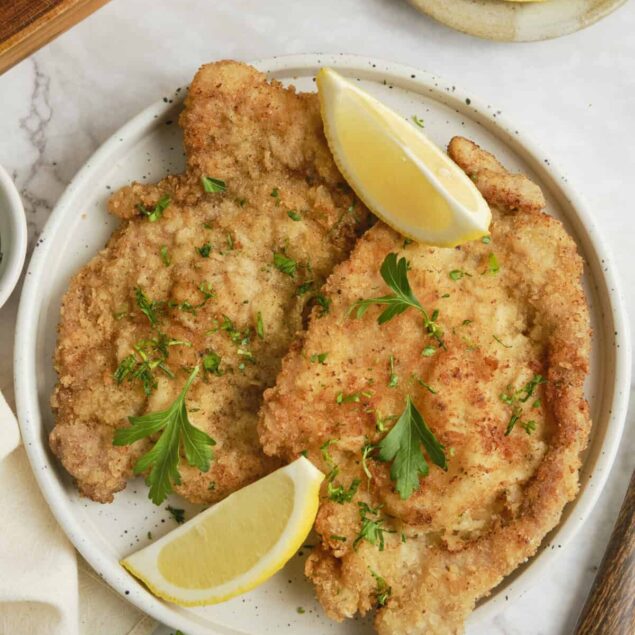 Golden brown German schnitzel on an elegant white plate, garnished with fresh lemon wedges and parsley, with a side of crispy potatoes