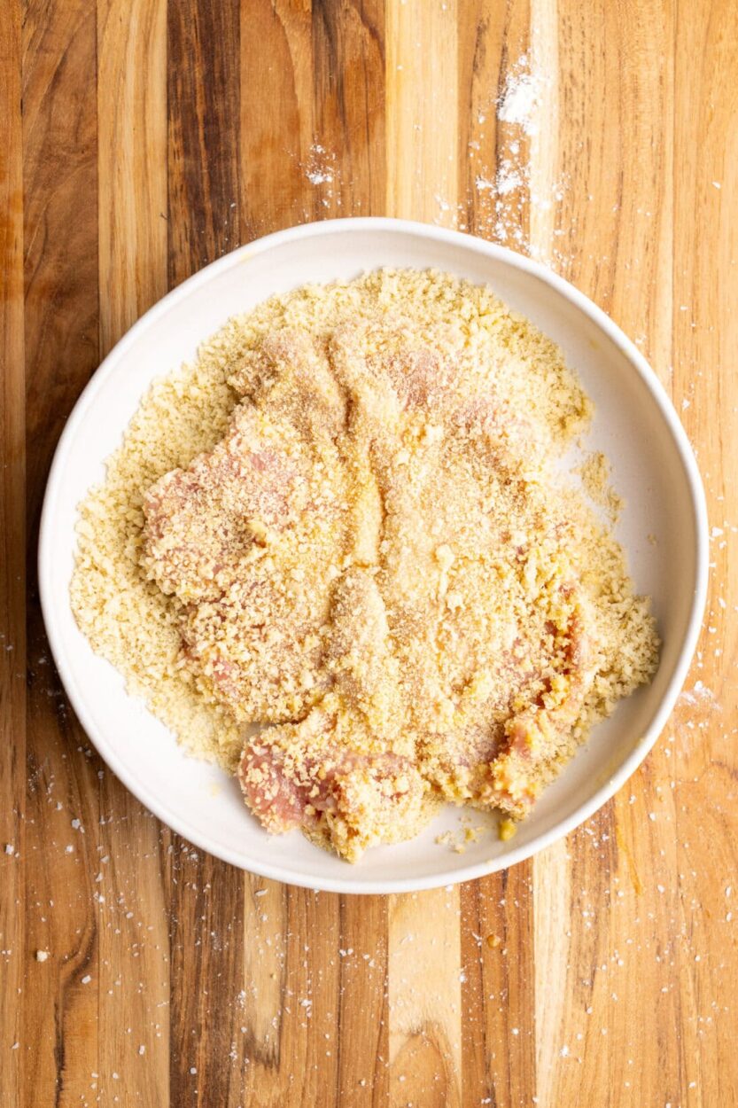 Three bowls showing the breading station setup - seasoned flour, beaten eggs with cream, and golden breadcrumbs, with pounded cutlets ready for coating