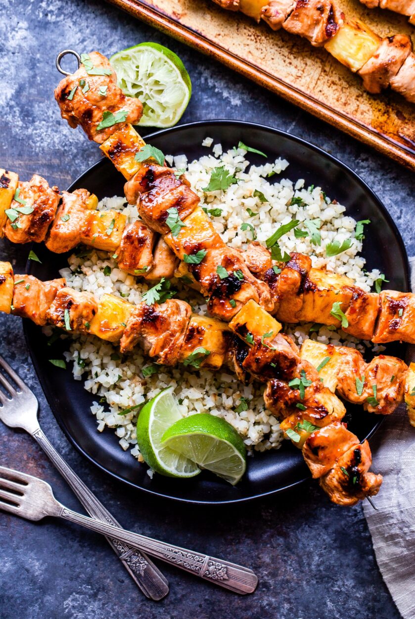 Close-up of grilled pineapple chicken skewer resting on a wooden serving board, showing caramelized char marks on pineapple and glistening glaze on chicken, garnished with fresh cilantro and lime wedges