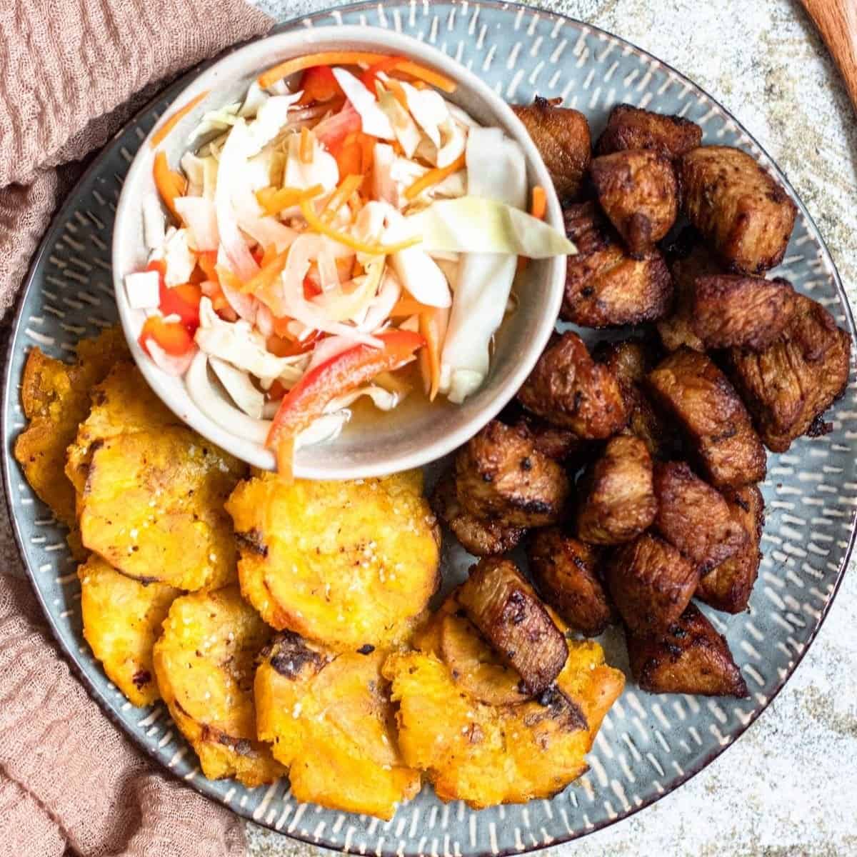 Close-up of crispy griot served over white rice with colorful pikliz and fried plantains on the side